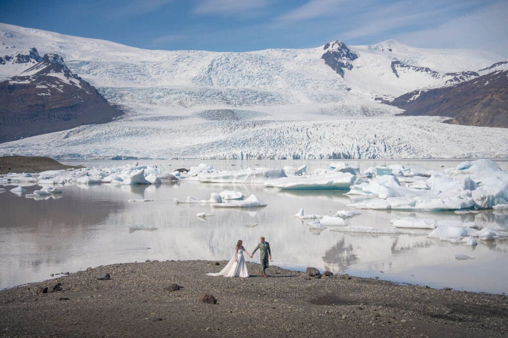 A couple in wedding attire is walking along the shore at Fjallsarlon glacier lagoon in Iceland