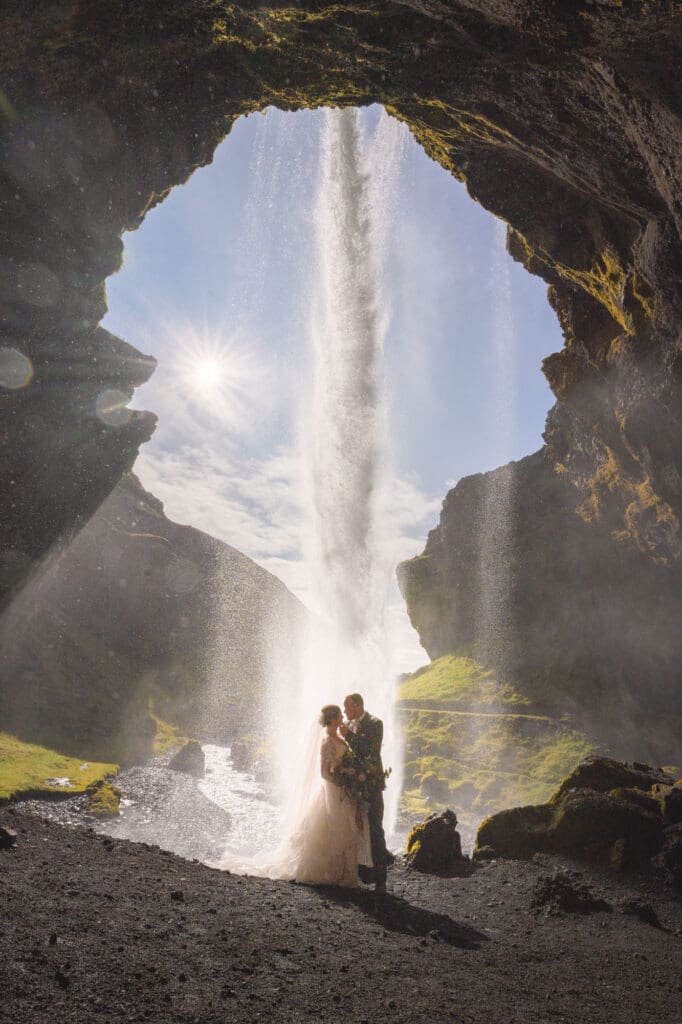 Couple in wedding attire standing behind backlit Kvernufoss waterfall in Iceland
