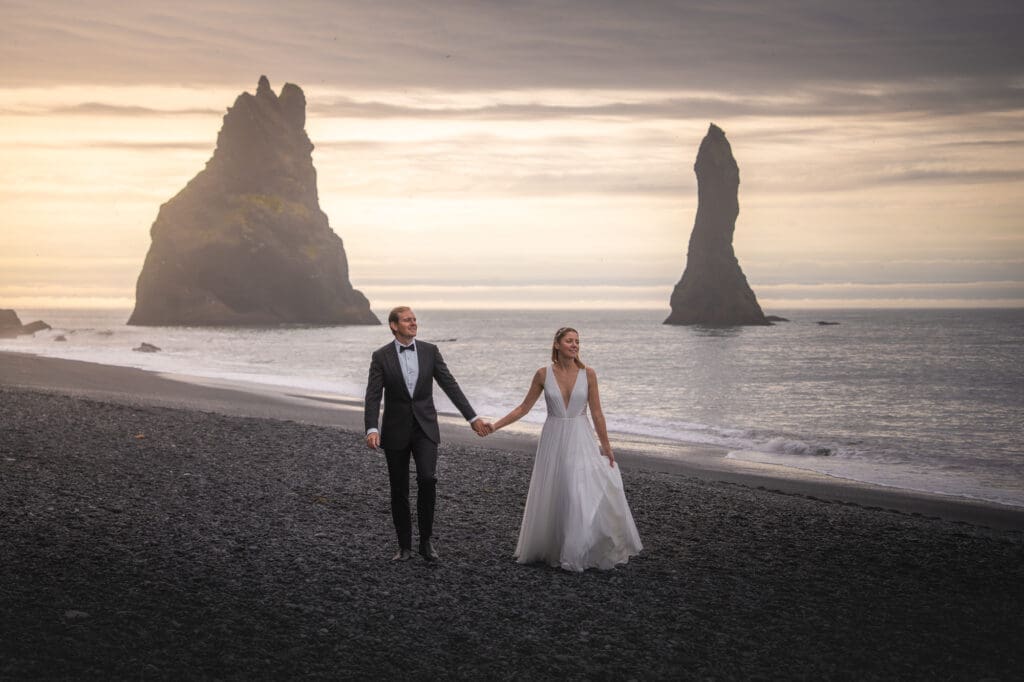 Couple in wedding attire is walking at Reynisfjara black sand beach between the sea stacks