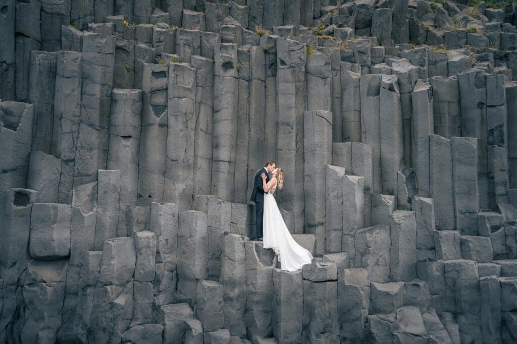 A couple in wedding attire are standing and kissing on basalt columns at Reynisfjara beach in Iceland