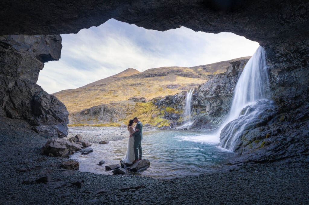 A couple in wedding attire is standing on a rock inside a cave next to a waterfall
