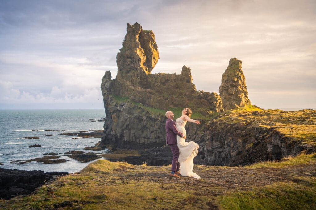 A groom is picking up his bride in front of a rock formation at sunset in Iceland 