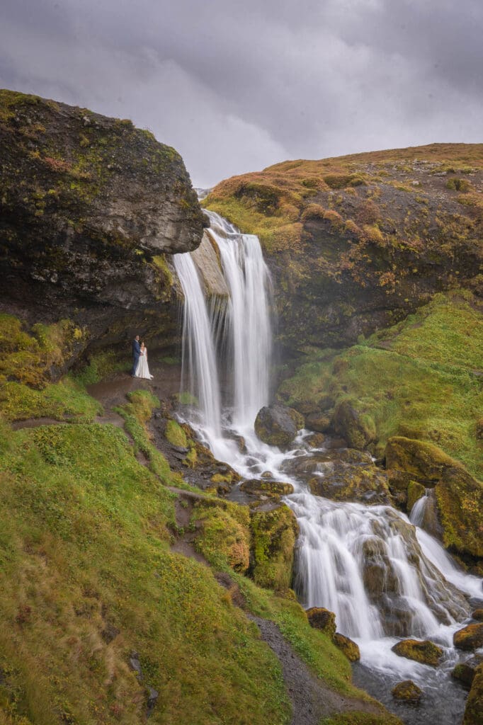 Bride and groom standing behind a smooth waterfall in Iceland