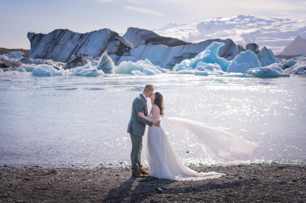 A couple in wedding attire are kissing in front on the shore of Jokulsarlon glacier lagoon in Iceland