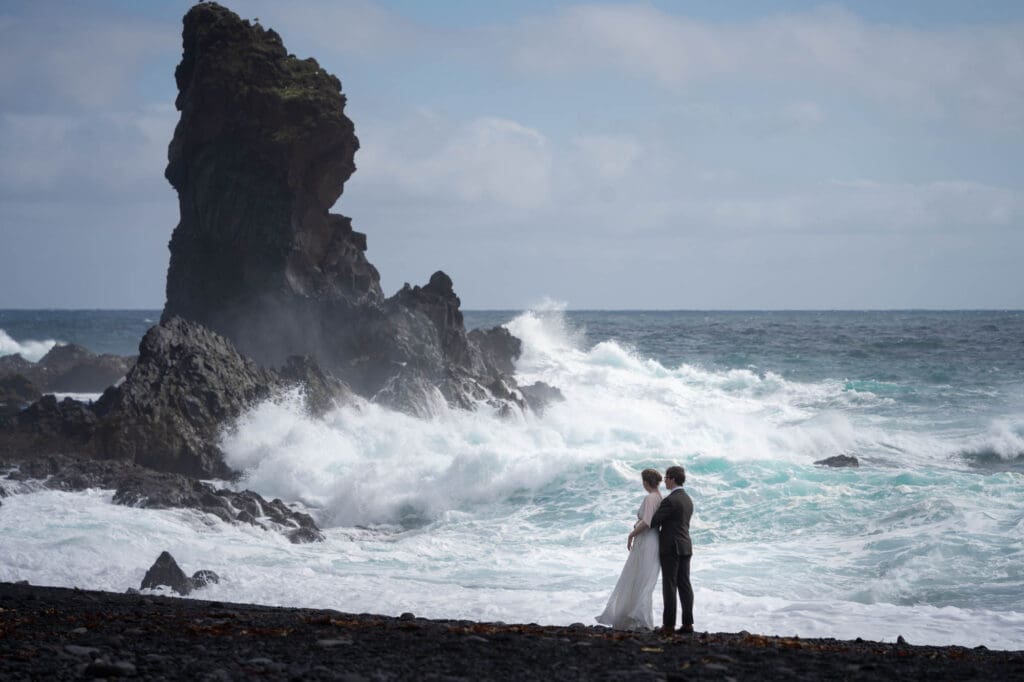A couple in wedding attire is standing on a black sand beach in Iceland and watching the waves