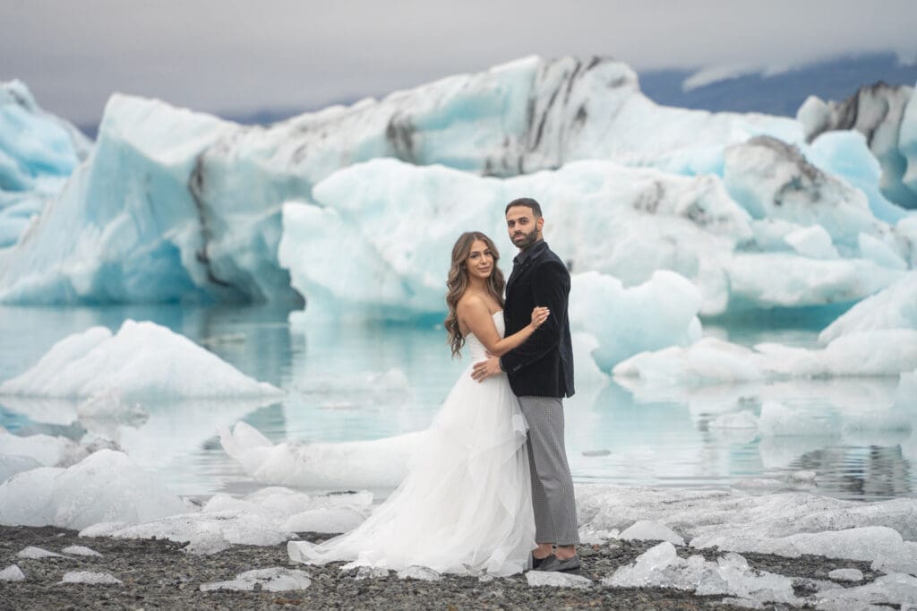 A couple in wedding attire is standing by a glacier lagoon full of large icebergs