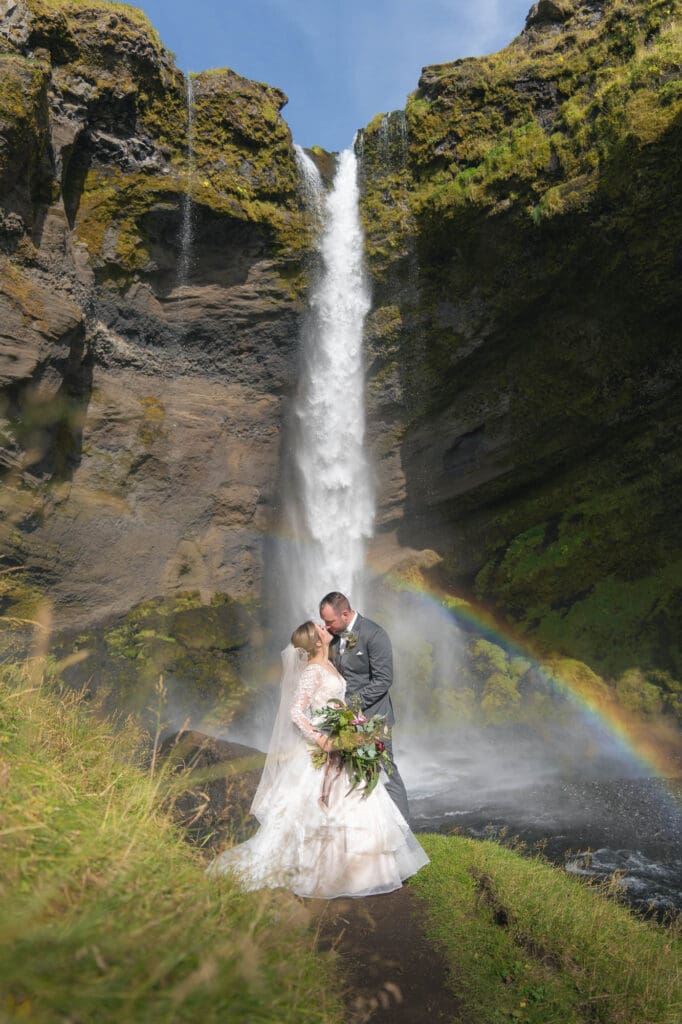 A couple in wedding attire is standing in front of Kvernufoss in Iceland with a rainbow behind them