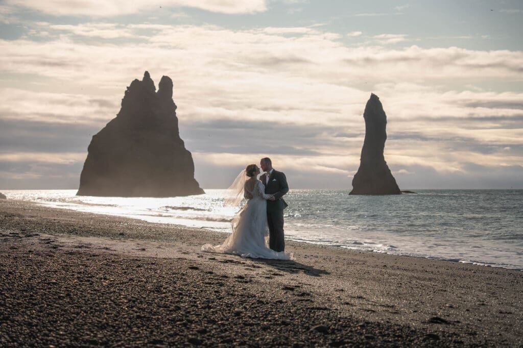 A couple in wedding attire is standing at Reynisfjara black sand between two sea stacks in Iceland