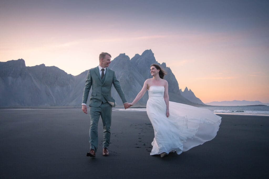 A couple in wedding attire is walking towards the camera. Vestrahorn mountain range is seen in the background