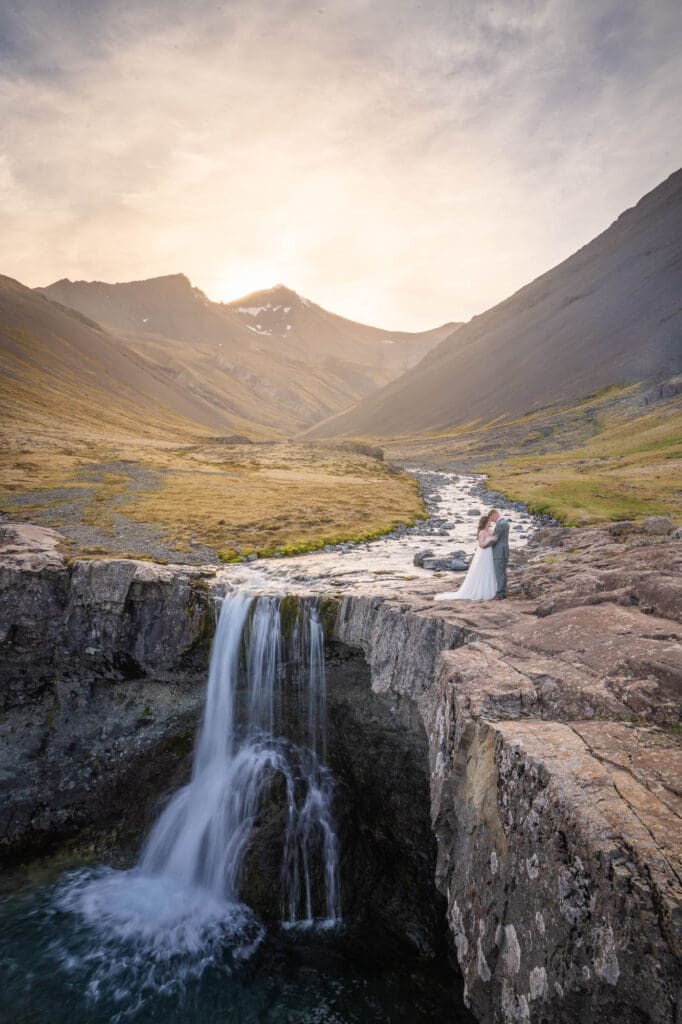 A couple in wedding attire is standing in front of a waterfall in Iceland at sunset