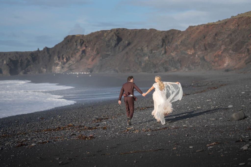 Couple in wedding attire is running at a blakc sand beach in Snaefellsnes Peninsula
