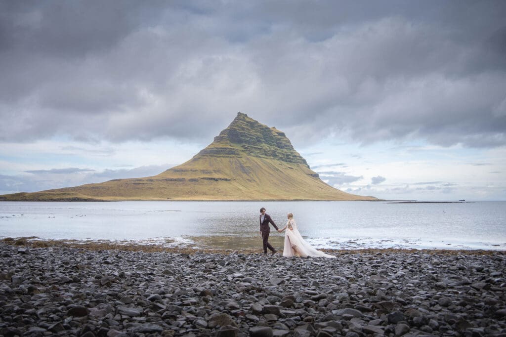 Couple in wedding attire is walking holding hands in front of Kirkjufell Mountain in Iceland