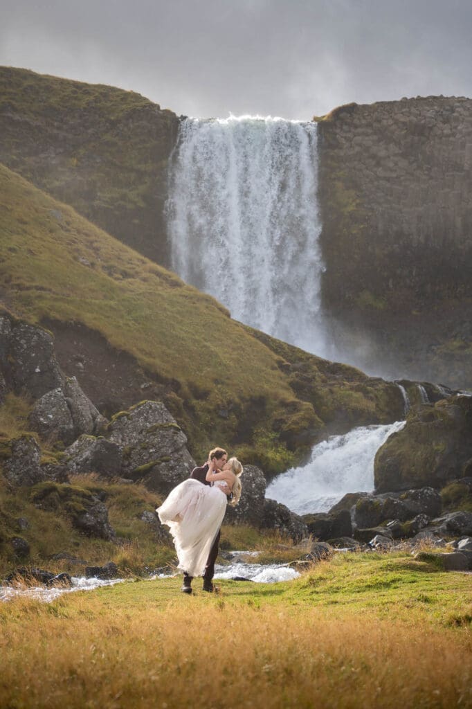 Groom is carrying his bride in front of a waterfall in Iceland